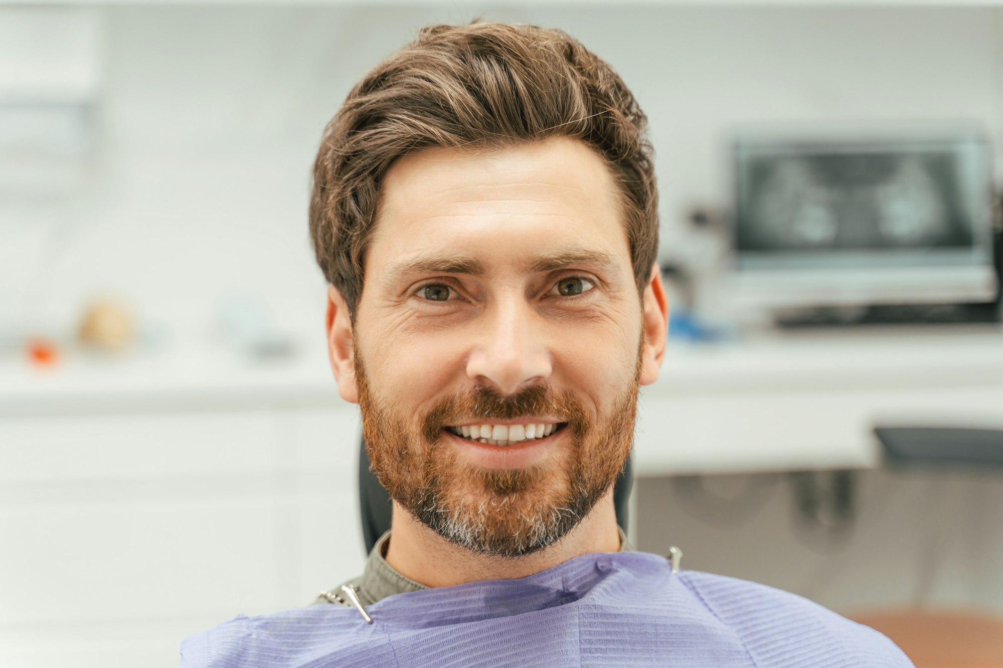 Smiling bearded man sitting in dental chair, looking at camera. Patient visiting dental clinic